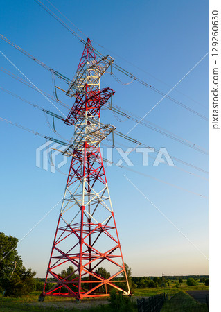 Red and white high-voltage transmission tower against blue sky in a rural landscape at sunset Red and white high-voltage transmission tower against blue sky in a rural landscape at sunset 129260630