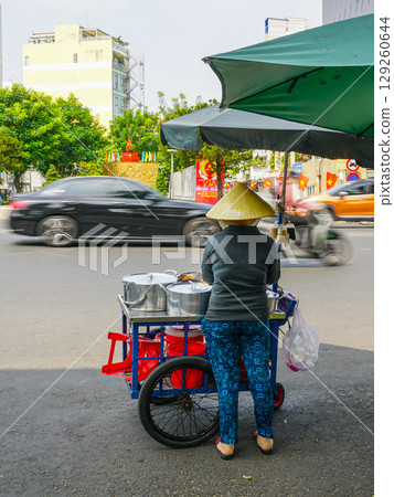 Vietnamese Street Food Vendor with Cart Under Umbrella as Traffic Flows on Busy Urban Road 129260644