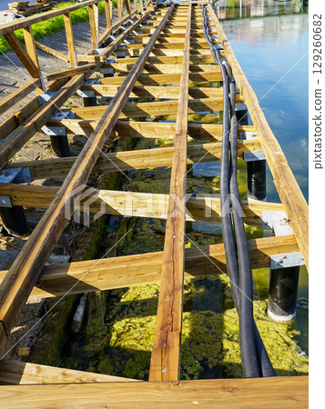 Wooden Pier Construction with Exposed Beams and Black Pipes over Shallow Algae-Filled Water Wooden Pier Construction with Exposed Beams and Black Pipes over Shallow Algae-Filled Water 129260682
