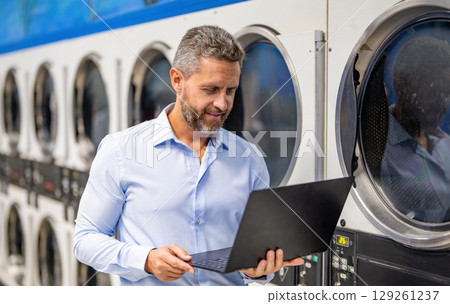 Busy man working on laptop at laundry. Overloaded employee multitasking laundry and job. Tired worker dealing laundry and deadlines. Laundry overload for busy businessman. Doing housework Busy man working on laptop at laundry. Overloaded employee multitasking laundry and job. Tired worker dealing laundry and deadlines. Laundry overload for busy businessman. Doing housework 129261237
