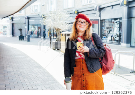 Young smiling plus size woman with backpack using mobile phone, drinking coffee outdoors on the street. Confident fashion hipster messaging, chatting, working remotely. Using navigator app for travel. 129262376