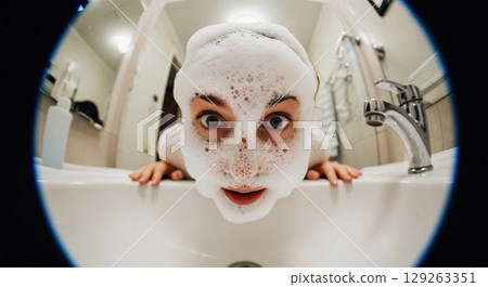 In a close-up view, a person leans over a sink, their face covered in frothy bubbles. The foam grows and pops around them, creating a playful atmosphere in a vibrant bathroom setting. 129263351