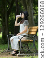 A young woman sitting on a bench in the shade of a tree and drinking water from her own bottle 129266068
