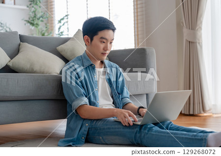 Young Asian man relaxing on comfortable couch, using laptop at home Young Asian man relaxing on comfortable couch, using laptop at home 129268072