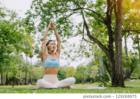 Asian woman meditating in a peaceful park setting, sitting cross-legged on green grass Asian woman meditating in a peaceful park setting, sitting cross-legged on green grass 129268075