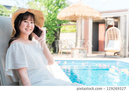 一位年輕的亞洲女性坐在泳池邊的躺椅上,用智慧型手機通話(預訂、搜尋、客房服務) 一位年輕的亞洲女性坐在泳池邊的躺椅上,用智慧型手機通話(預訂、搜尋、客房服務) 129269327