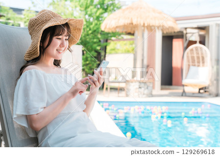 A young Asian woman sitting on a recliner by the pool and operating her smartphone 129269618