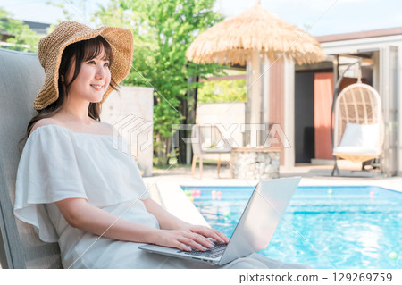 A young Asian woman sitting on a recliner by the pool and using a laptop 129269759