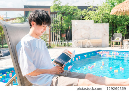 一位年輕的亞洲男子坐在泳池邊的躺椅上閱讀旅遊指南 129269869