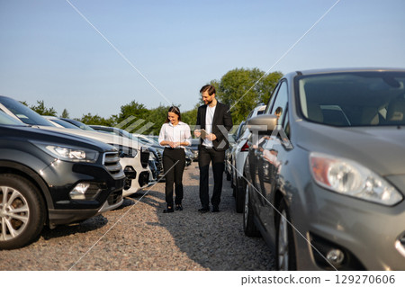 A male car dealer assisting a female customer as they walk through a car lot, possibly discussing vehicle options. 129270606
