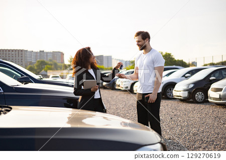 A female car dealer hands car keys to a male customer in a car dealership lot under sunlight. A female car dealer hands car keys to a male customer in a car dealership lot under sunlight. 129270619