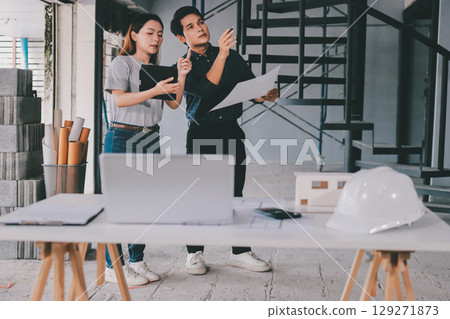 Construction manager and engineer dressed in orange work vests and hard helmets explore construction documentation on the building site near the steel frames 129271873