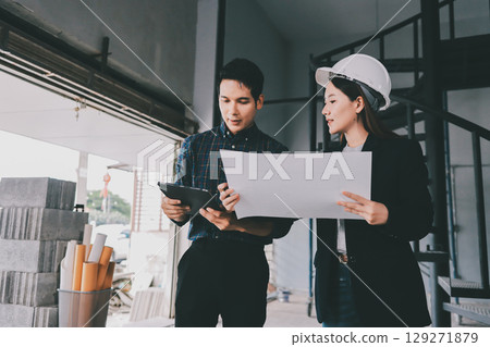 Construction manager and engineer dressed in orange work vests and hard helmets explore construction documentation on the building site near the steel frames 129271879