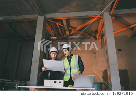 Construction manager and engineer dressed in orange work vests and hard helmets explore construction documentation on the building site near the steel frames Construction manager and engineer dressed in orange work vests and hard helmets explore construction documentation on the building site near the steel frames 129271910