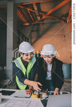 Construction manager and engineer dressed in orange work vests and hard helmets explore construction documentation on the building site near the steel frames 129271918