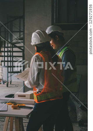 Construction manager and engineer dressed in orange work vests and hard helmets explore construction documentation on the building site near the steel frames Construction manager and engineer dressed in orange work vests and hard helmets explore construction documentation on the building site near the steel frames 129271936