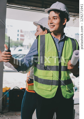 Construction manager and engineer dressed in orange work vests and hard helmets explore construction documentation on the building site near the steel frames Construction manager and engineer dressed in orange work vests and hard helmets explore construction documentation on the building site near the steel frames 129271941