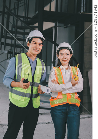 Construction manager and engineer dressed in orange work vests and hard helmets explore construction documentation on the building site near the steel frames 129271942