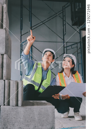 Construction manager and engineer dressed in orange work vests and hard helmets explore construction documentation on the building site near the steel frames 129271946
