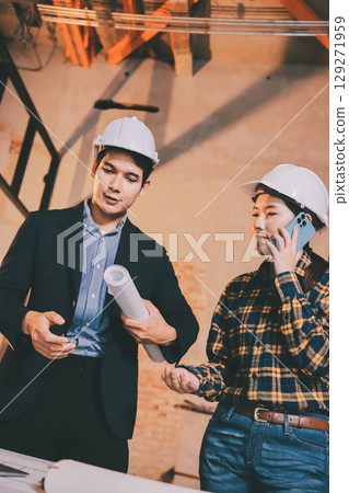 Construction manager and engineer dressed in orange work vests and hard helmets explore construction documentation on the building site near the steel frames Construction manager and engineer dressed in orange work vests and hard helmets explore construction documentation on the building site near the steel frames 129271959