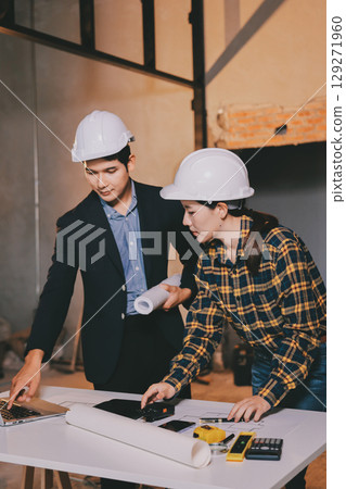 Construction manager and engineer dressed in orange work vests and hard helmets explore construction documentation on the building site near the steel frames Construction manager and engineer dressed in orange work vests and hard helmets explore construction documentation on the building site near the steel frames 129271960