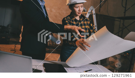 Construction manager and engineer dressed in orange work vests and hard helmets explore construction documentation on the building site near the steel frames Construction manager and engineer dressed in orange work vests and hard helmets explore construction documentation on the building site near the steel frames 129271961