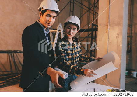 Construction manager and engineer dressed in orange work vests and hard helmets explore construction documentation on the building site near the steel frames 129271962