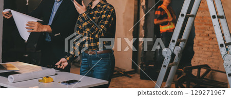 Construction manager and engineer dressed in orange work vests and hard helmets explore construction documentation on the building site near the steel frames Construction manager and engineer dressed in orange work vests and hard helmets explore construction documentation on the building site near the steel frames 129271967