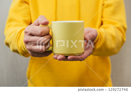 person in yellow hoodie holds yellow mug. cozy moment indoors. copy space. closeup. 129272748