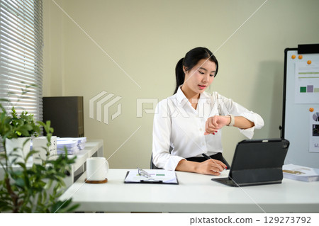 Businesswoman checking time on smartwatch while working on tablet at a modern office desk 129273792