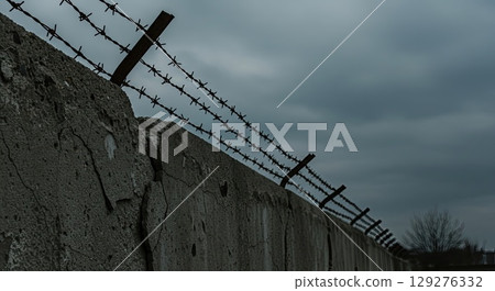 Concrete wall with barbed wire under gloomy sky symbolizes historical oppression 129276332