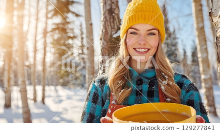 A smiling woman in a yellow hat enjoys the winter forest, holding a bucket, surrounded by snow and sunlight 129277841