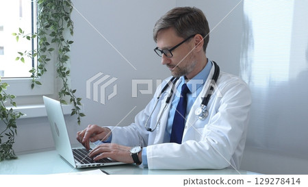 Male doctor wearing a lab coat and stethoscope, sitting at a desk and typing on a laptop computer in a modern medical office, focused on patient care and consultation. Medicine and health care Male doctor wearing a lab coat and stethoscope, sitting at a desk and typing on a laptop computer in a modern medical office, focused on patient care and consultation. Medicine and health care 129278414