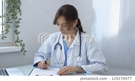Female doctor in lab coat and stethoscope diligently writing notes on clipboard while sitting at table in medical office with window. Medicine concept. Female doctor in lab coat and stethoscope diligently writing notes on clipboard while sitting at table in medical office with window. Medicine concept. 129278477