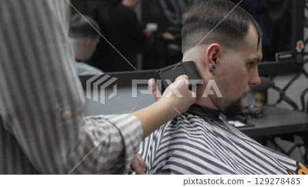 Professional barber trimming customer's hair with electric clipper, focusing on precise nape grooming within contemporary barbershop interior design 129278485