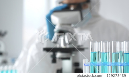 Medical scientist wearing protective suit using microscope while analyzing samples, conducting research in laboratory environment. Close up of test tubes with blue liquid 129278489