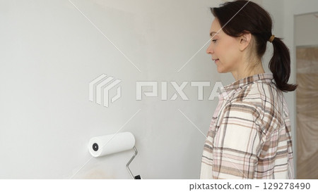 Painter woman wearing gloves is skillfully applying paint to a white wall with a roller in an apartment undergoing renovation, transforming the space with her creative touch 129278490
