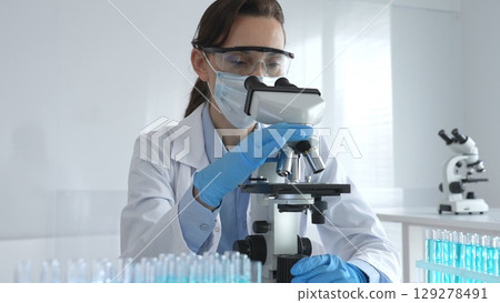 Female scientist in lab coat and safety glasses using microscope, analyzing samples in test tubes in laboratory, conducting scientific research Female scientist in lab coat and safety glasses using microscope, analyzing samples in test tubes in laboratory, conducting scientific research 129278491
