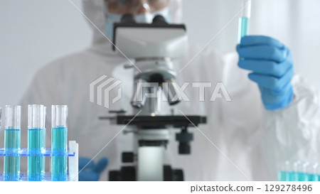 Medical scientist in protective suit and gloves using microscope while analyzing samples, conducting research in laboratory. Close-up of test tubes with blue liquid Medical scientist in protective suit and gloves using microscope while analyzing samples, conducting research in laboratory. Close-up of test tubes with blue liquid 129278496