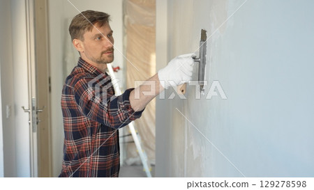 Male construction worker wearing gloves is carefully applying plaster to a wall using a hand tool, contributing to a home renovation or construction project Male construction worker wearing gloves is carefully applying plaster to a wall using a hand tool, contributing to a home renovation or construction project 129278598