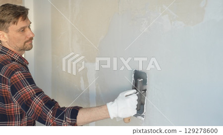 Male construction worker wearing gloves is carefully applying plaster to a wall using a hand tool, contributing to a home renovation or construction project Male construction worker wearing gloves is carefully applying plaster to a wall using a hand tool, contributing to a home renovation or construction project 129278600