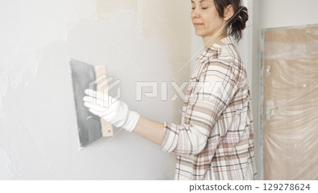 Woman applying plaster to wall with putty knife, wearing protective gloves and creating smooth surface during home renovation work 129278624