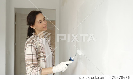Middle aged woman wearing protective gloves painting white wall with paint roller, updating interior and giving room clean, modern look during home renovation 129278626