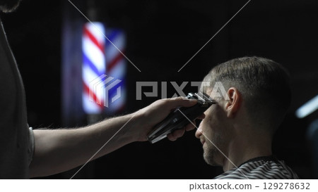 Skilled barber trimming client's hair using electric clipper, vintage barbershop interior with blurred pole, professional grooming service 129278632