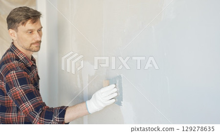 Construction worker man wearing gloves and holding a putty knife is applying plaster on a white wall, performing home renovation and improvement 129278635