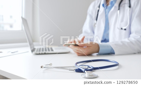 Closeup of blue stethoscope is lying on the table near female doctor checking smartphone representing digital healthcare workflow in well lit clinical workspace. Medicine and health care Closeup of blue stethoscope is lying on the table near female doctor checking smartphone representing digital healthcare workflow in well lit clinical workspace. Medicine and health care 129278650