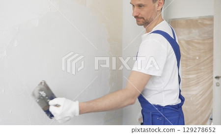 Construction worker plastering white wall, wearing blue overalls and protective white gloves during apartment renovation project 129278652