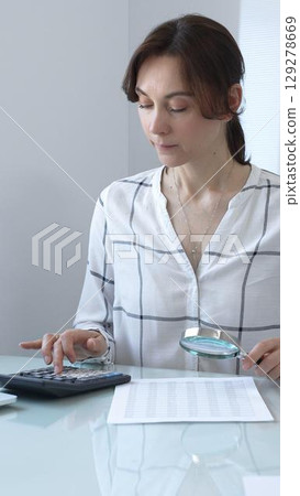 Accountant carefully examines financial documents, enlarges details with a magnifying glass, calculates performance indicators in a professional workplace. Audit and taxes in business. 129278669