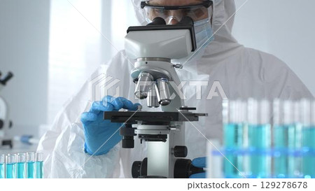 A male doctor in a protective suit and gloves uses a microscope while analyzing samples, conducting research in a laboratory. Close-up of test tubes with blue liquid 129278678