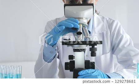 A female scientist in protective gear adjusts a microscope lens and accurately analyzes medical research samples in a sterile laboratory setting 129278681
